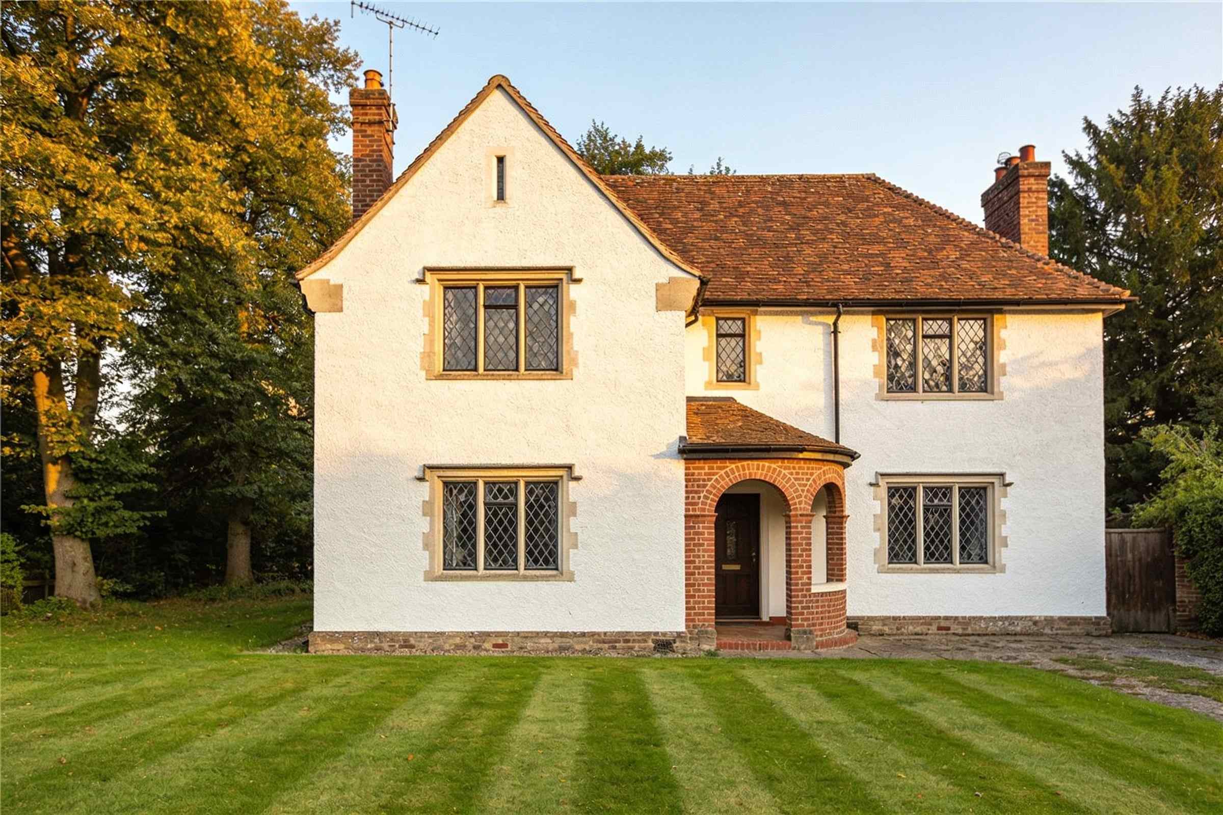 A white-rendered 1930s arts and crafts house with a steep clay tiled roof and a long lawn, in late summer evening light.