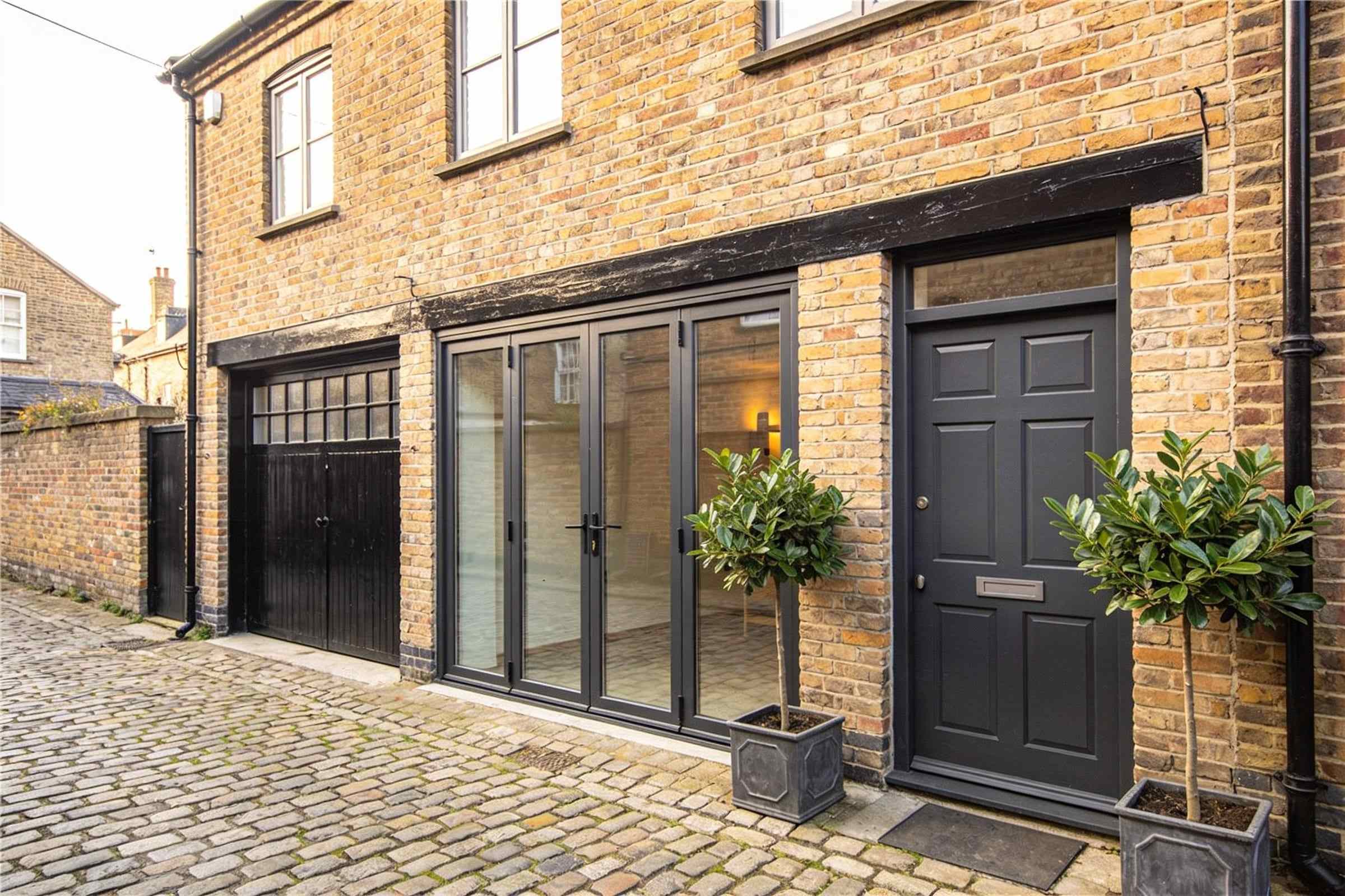 A brick mews house with glazed doors, a dark grey front door and potted bay trees on a cobbled lane.