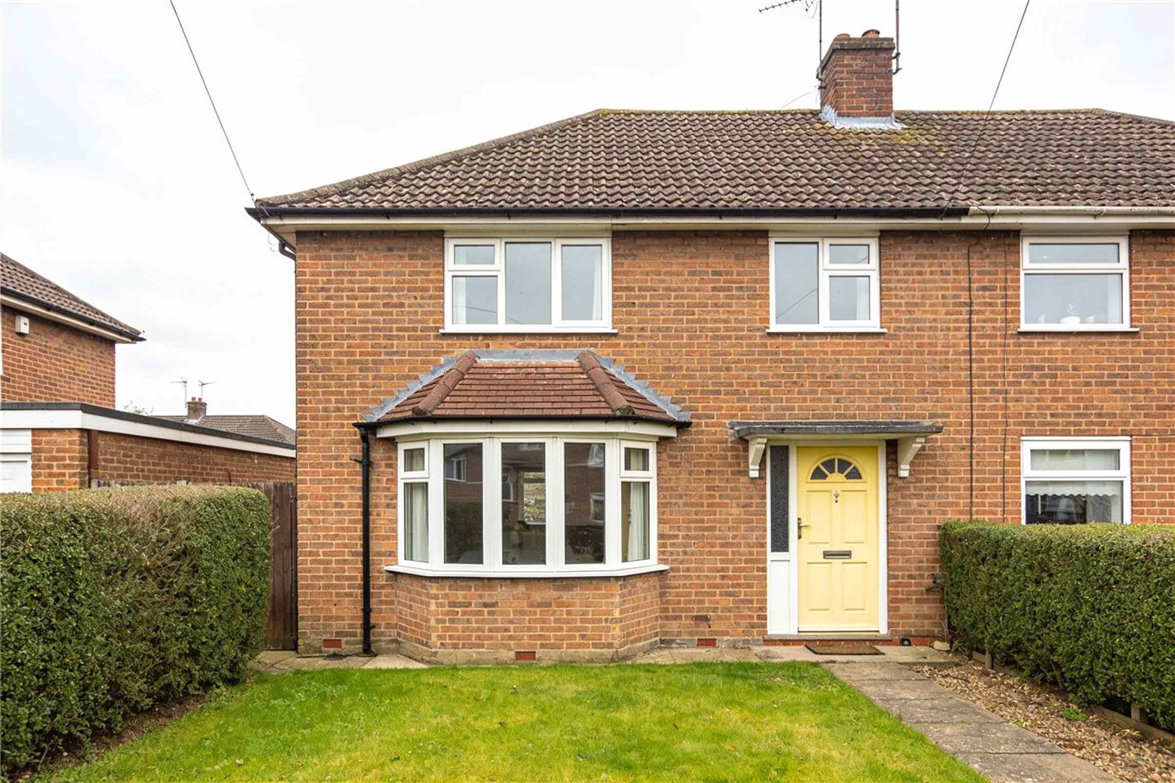 A brick 1950s semi-detached house with a bay window, a soft yellow front door and a front lawn behind a hedge.