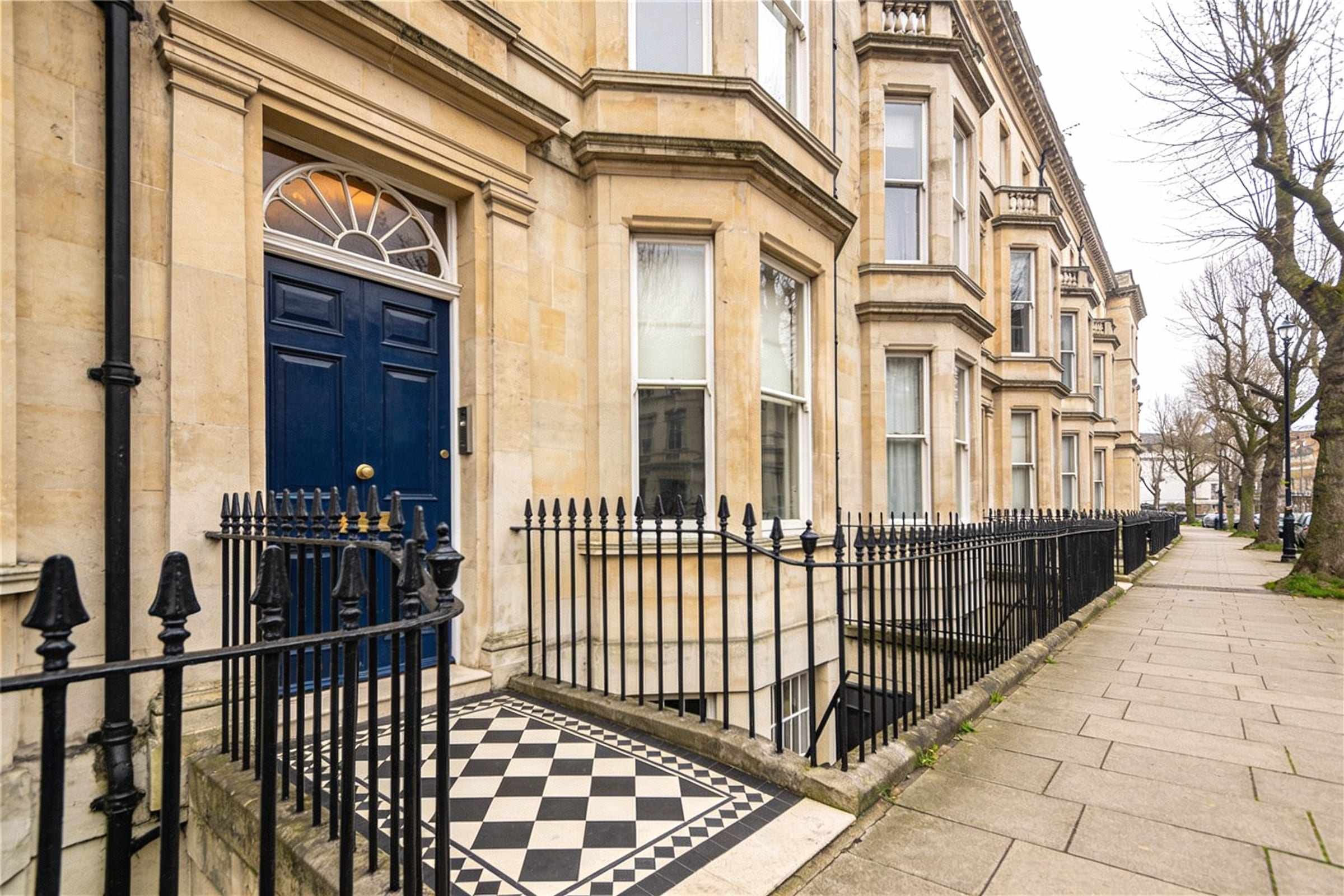 A tall Victorian stone townhouse with a blue front door, black railings and a tiled path on a tree-lined street.
