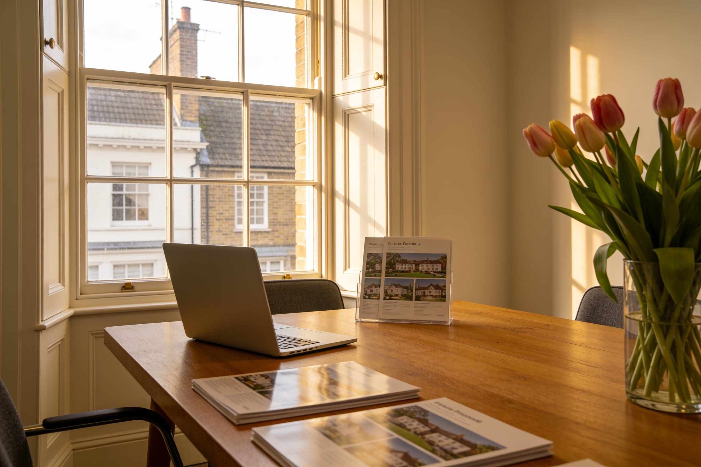 A meeting table by a tall sash window with a laptop, brochures and a vase of tulips.