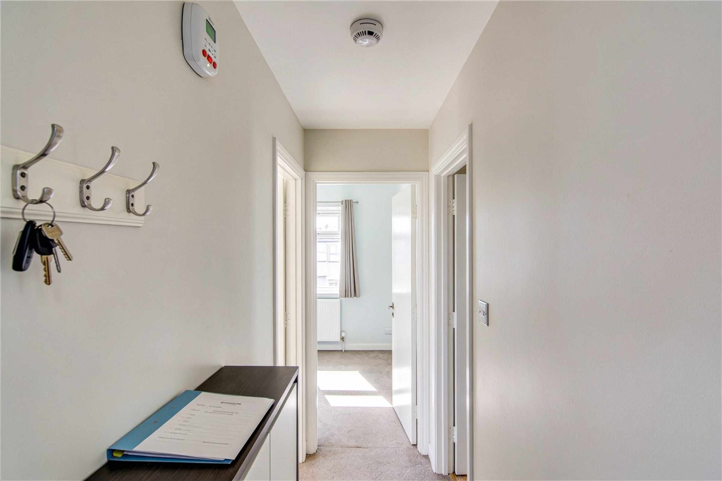 A tidy rented hallway with a smoke alarm on the ceiling, keys on a hook and a folder on a console table.