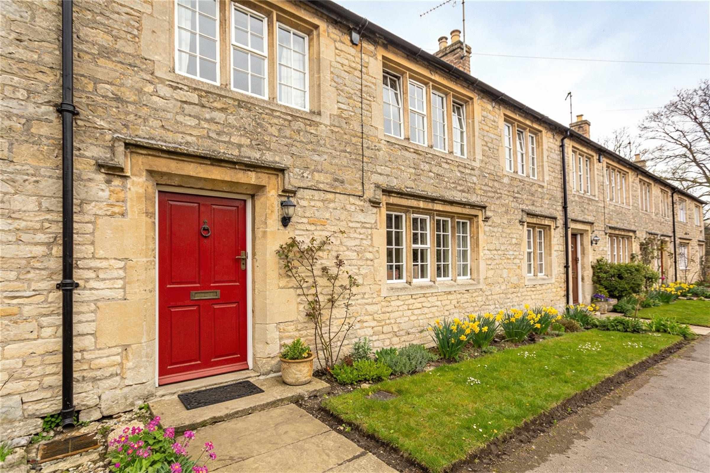 A stone weaver’s cottage with long upper windows and a red front door in a terrace of cottages, in spring light.