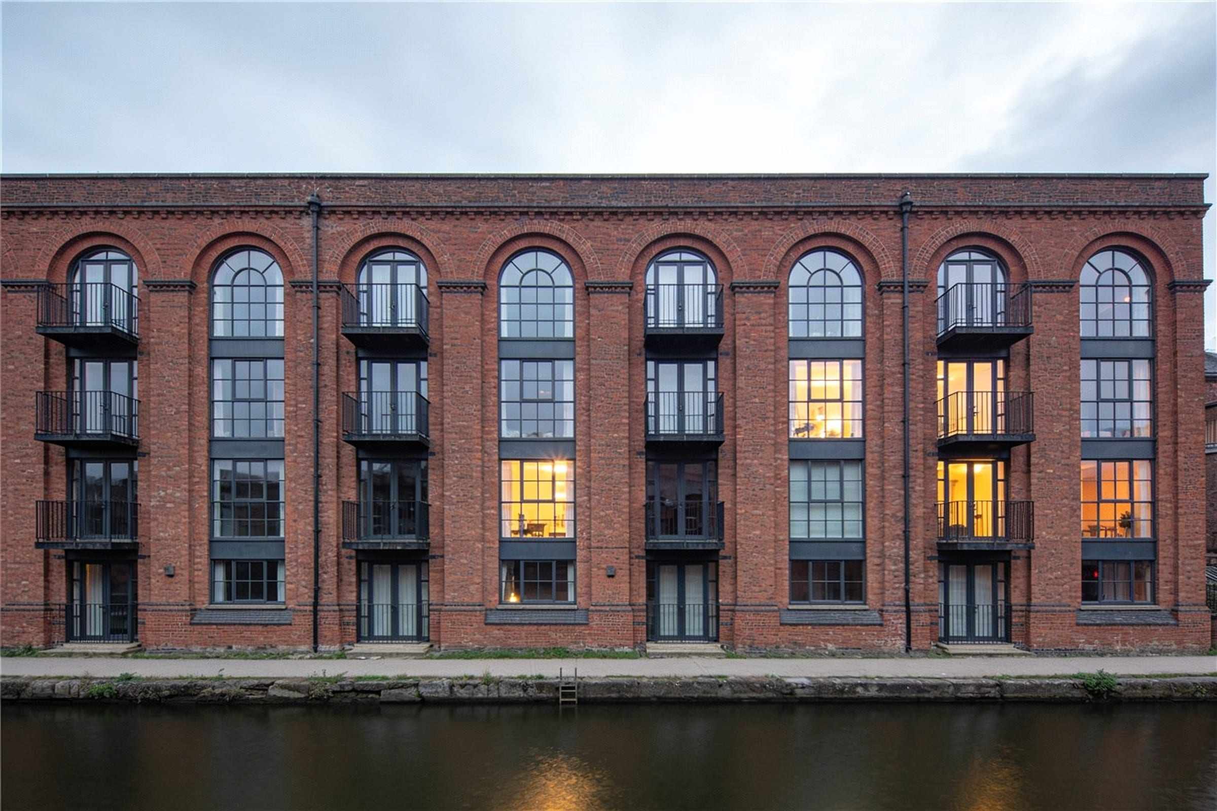 A converted red-brick warehouse with tall arched windows and small balconies on the river, at dusk.