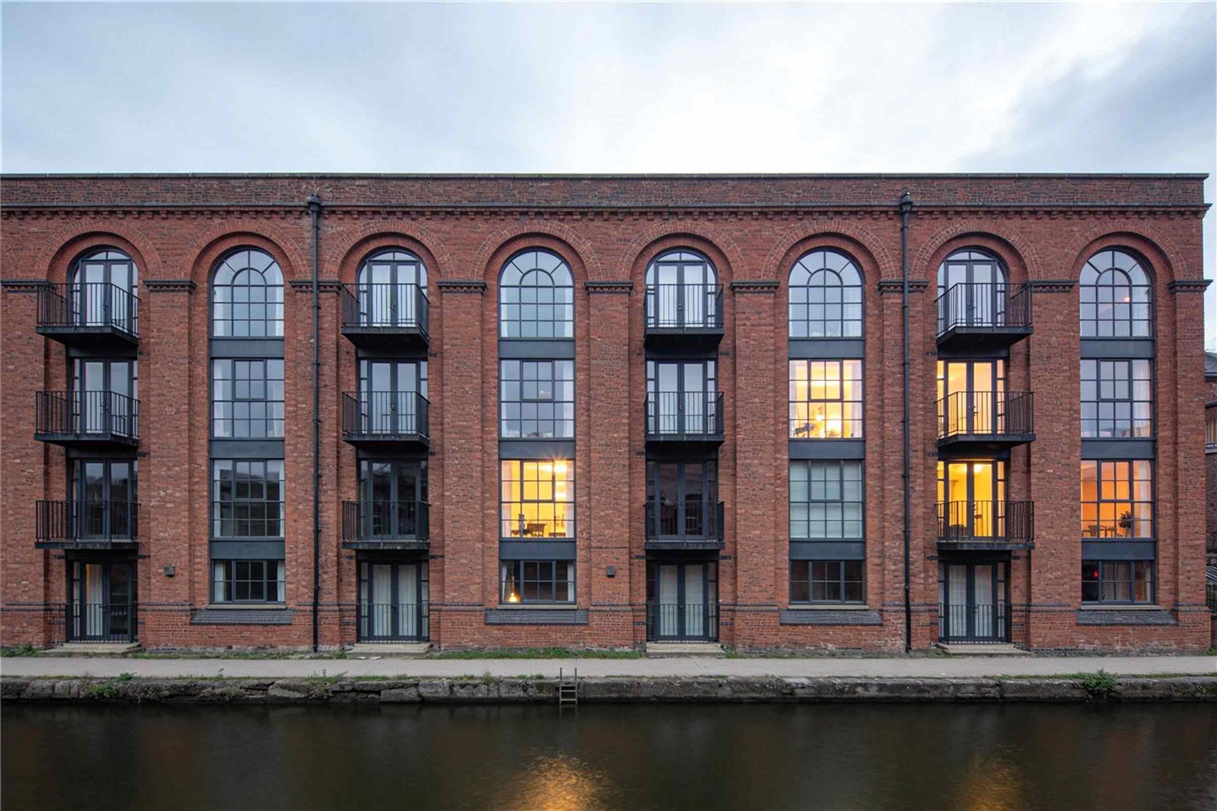 A converted red-brick warehouse with tall arched windows and small balconies on the river, at dusk.